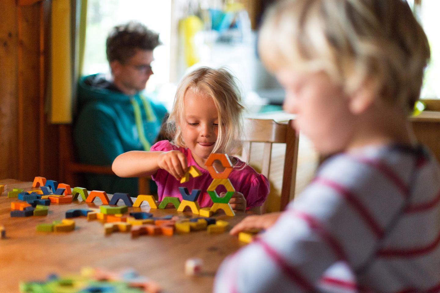 Spielzeug ab 5 Jahren: Mädchen am Küchentisch mit Bruder stapelt konzentriert bunte Bauklötze.