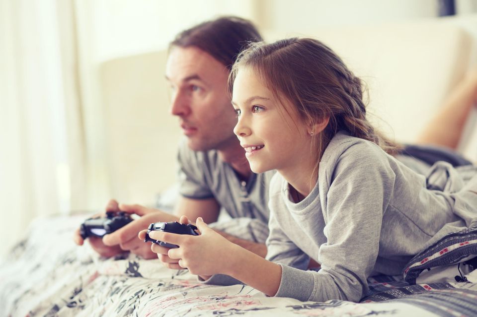 Spielekonsolen für Kinder: Mädchen spielt mit ihrem Papa gemeinsam an der Playstation.