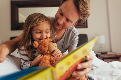 Kinderbücher ab 3 Jahren: Lachendes Mädchen mit Teddybär schaut mit ihrem Vater ein Buch an.