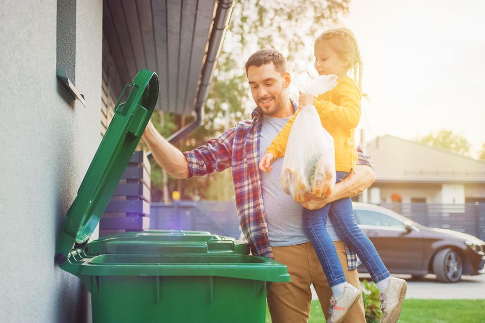 Familienkolumne: Ein Vater hat seine Tochter auf dem Arm und öffnet einen Mülltonnendeckel