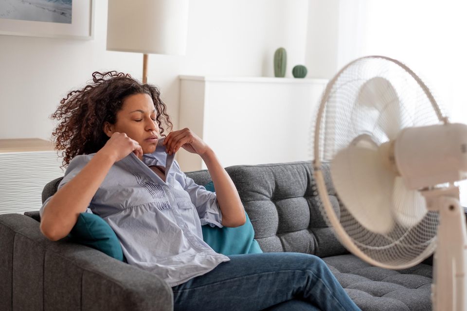 Hitzewallungen in der Schwangerschaft: Frau mit blauer Bluse sitzt auf dem Sofa vor einem laufenden Ventilator