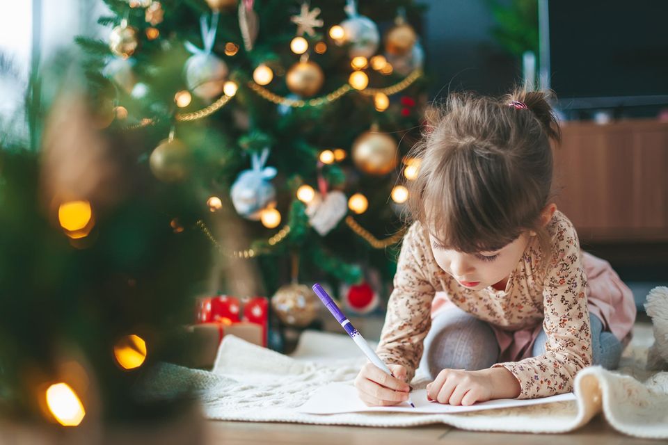 Lichterketten ja oder nein?: Kind sitzt vor beleuchtetem Weihnachtsbaum und schreibt ihren Wunschzettel