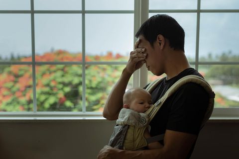 Elternsein: Ein gestresster Vater steht mit seinem Baby in einer Trage am Fenster
