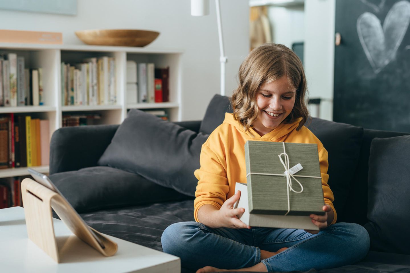 Geschenke für Teenager: Glücklich lächelndes Teeniemädchen mit geöffneter Geschenkebox auf dem Sofa.