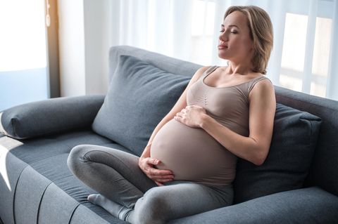Hochschwangere Frau mit geschlossenen Augen im Schneidersitz auf dem Sofa