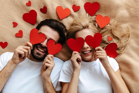 Zum Valentinstag: Mann und Frau liegen auf dem Rücken umgeben von Herzen und halten sich die Augen mit Herzen zu