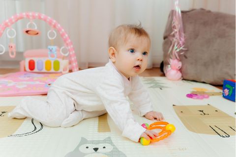Babyspielzeug ab 6 Monate: Baby im weißen Strampler in Krabbelstellung mit Spielsachen auf Spielmatte.