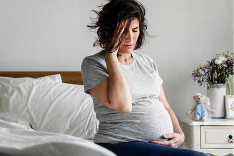 Ibuprofen Schwangerschaft: Schwangere sitzt auf dem Bett und hält sich den schmerzenden Kopf