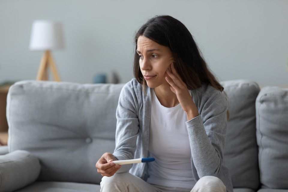 Alles-oder-nichts-Prinzip: Frau sitzt mit einem Schwangerschaftstest nachdenklich auf dem Sofa