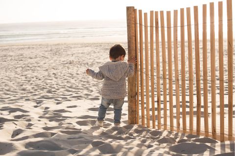 Sonnencreme für Babys: Kleines Kind steht am Strand und hält sich an Holzzaun fest