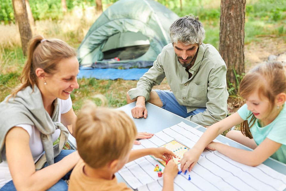 Reisespiele: Eltern mit zwei Kindern spielen "Mensch ärgere dich nicht" auf dem Campingplatz.
