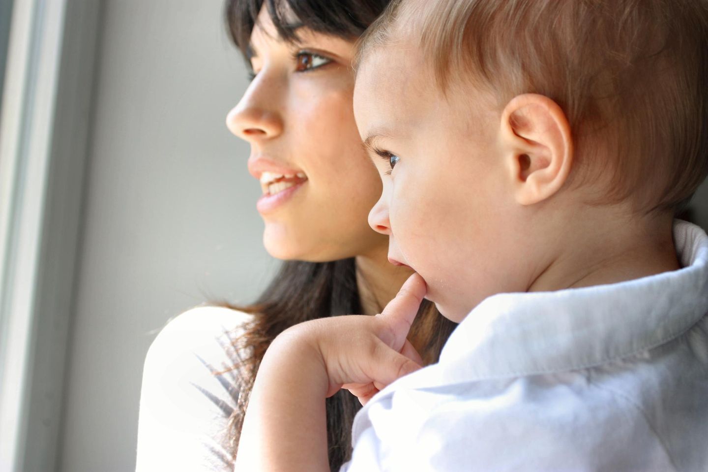 Frau mit Kleinkind auf dem Arm, beide schauen in die gleiche Richtung, das Kind mit Finger im Mund