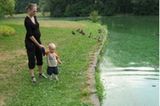 Schwangere Frau mit Kind an der Hand steht an einem Gewässer. Im Hintergrund Enten.