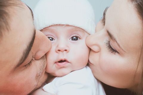 Was liebt ihr an eurem Baby am meisten: Eltern küssen Baby mit weißem Mützchen von rechts und links auf die Wange