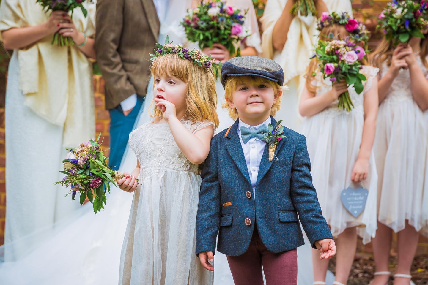 Festliche Kindermode: Mädchen und Junge, festlich gekleidet, auf einer Hochzeit mit Blumenmädchen im Hintergrund.