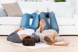 Mutmacher in der Kinderwunschzeit: Paar liegt Händchen haltend auf dem Fußboden, die Beine auf dem Sofa