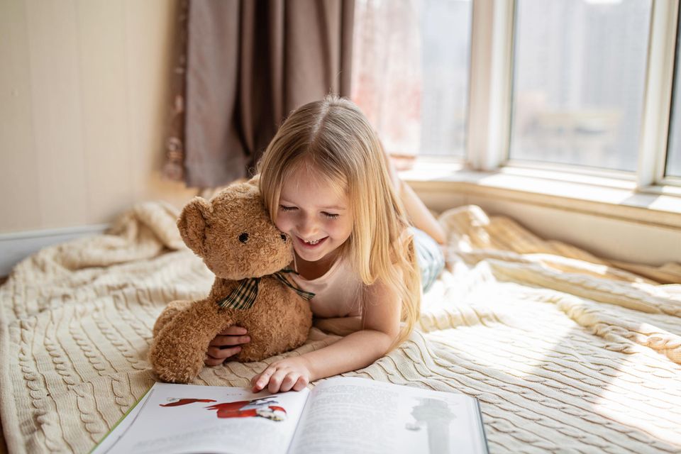 Zahnfee-Bücher: Mädchen mit Zahnlücke liegt mit Teddy auf Decke und schaut ein Buch an.