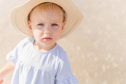 Hitzschlag bei Kindern: Blondes Kleinkind mit Strohhut auf dem Kopf blinzelt in die Sonne
