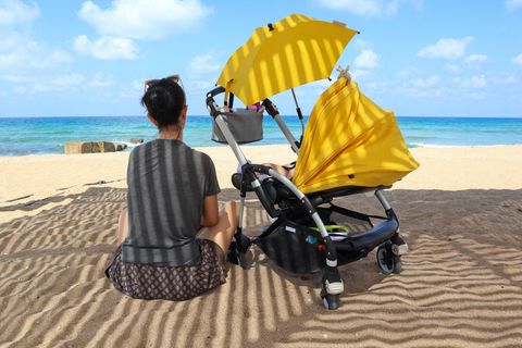 Sonnenschirm für Kinderwagen: Mutter sitzt neben Buggy mit Sonnenschirm am Strand.