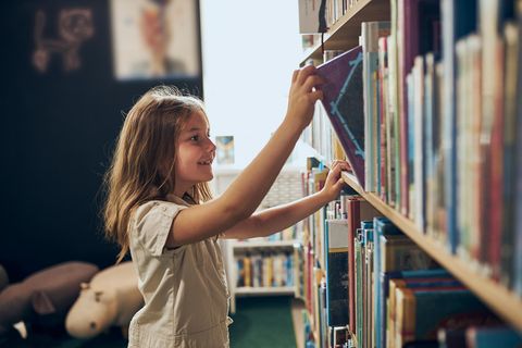 Ein Mädchen zieht sich in einer Bibliothek freudig ein buch aus dem Regal