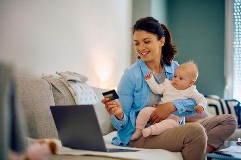 Angebote für Eltern: Mutter mit Baby auf dem Arm shoppt mit Kreditkarte vor dem Laptop
