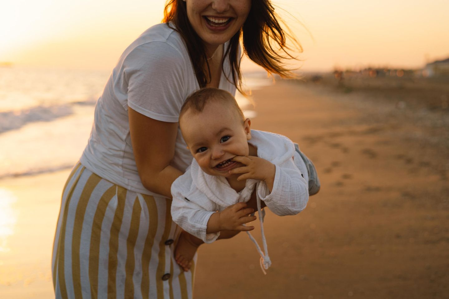 Reisen in der Elternzeit: Mutter hält lachendes Baby im Fliegergriff, Abendstimmung am Strand