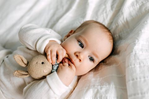 Babyspielzeug für 3 Monate altes Kind: Baby in Weiß beißt auf einer Holzrassel mit Häkelhase rum.