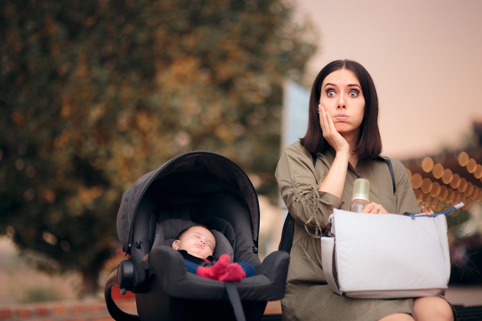 Erstaunte Mutter mit Wickeltasche und Baby in der Autoschale auf Parkbank