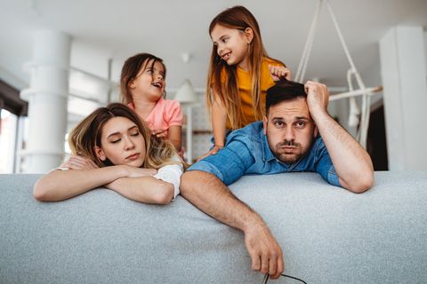 Burnout-Gefahr? Mann und Frau erschöpft auf Sofa, zwei Kindern turnen auf ihnen herum