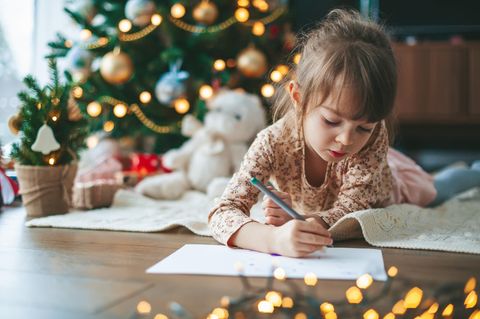 Kind liegt auf dem Boden vor einem Weihnachtsbaum und schreibt einen Brief