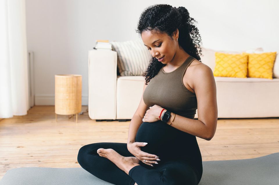 Yoga für Schwangere: Junge schwarze Frau im Schneidersitz mit beiden Händen auf ihrem Babybauch