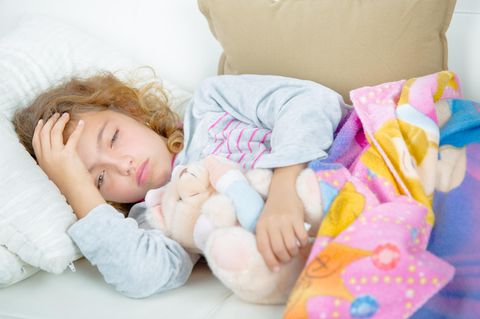 Kopfschmerzen bei Kindern: Kleines Mädchen liegt im Bett, hält sich den Kopf und wirkt krank