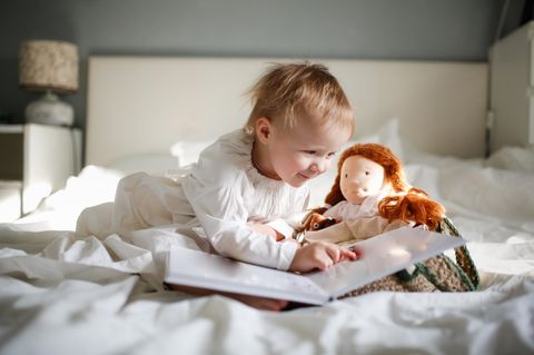 Puppen-Test: Kleinkind schaut mit Stoffpuppe ein Buch auf dem Bett an.