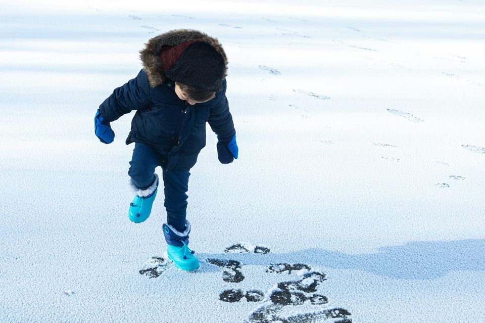 Kinderschuhe im Winter: Warm angezogenes Kind macht mit Schneestiefeln Fußspuren in den Schnee.