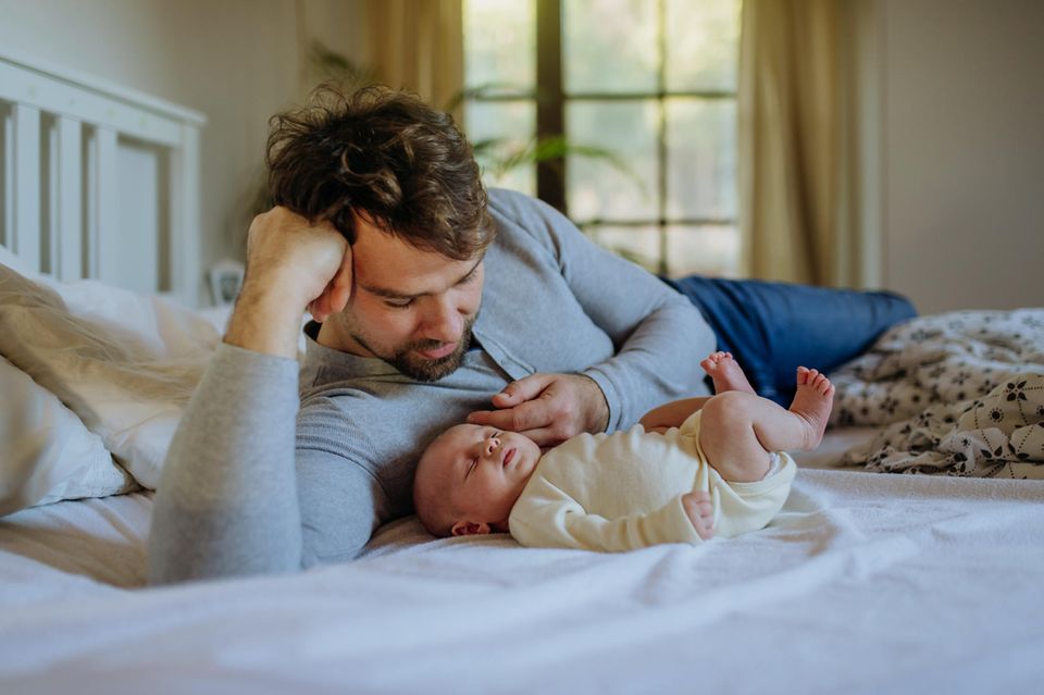 Vaterschaftsurlaub 2024: Mann liegt mit neugeborenem Baby auf dem Bett