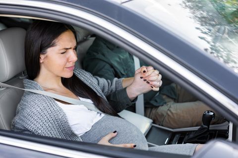 Schwangere Frau sitzt als Beifahrerin in einem Auto und hält die Hand der fahrenden Person