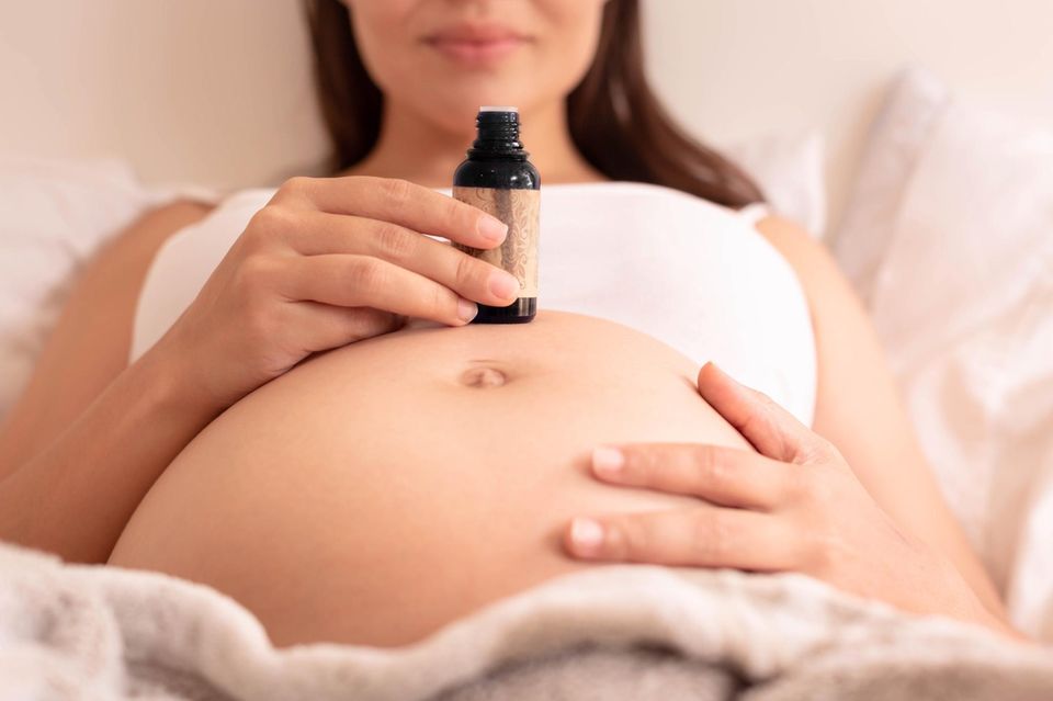 Dammmassage: Liegende Schwangere stellt eine Flasche mit Massageöl auf ihrem Bauch ab