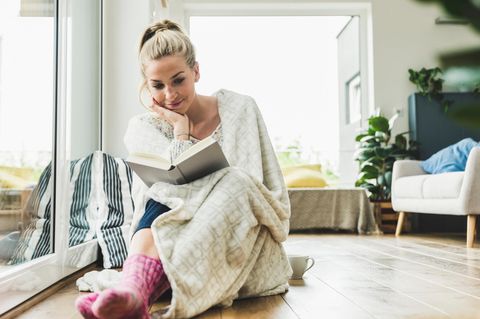 Junge Frau mit Decke liest schmunzelnd ein Buch auf dem Fußboden.
