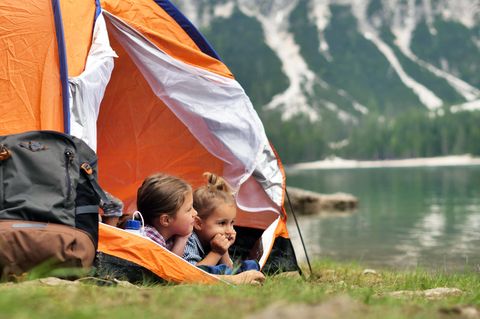 Camping mit Kindern: Zwei Mädchen schauen vor Berglandschaft aus einem Zelt am See heraus.
