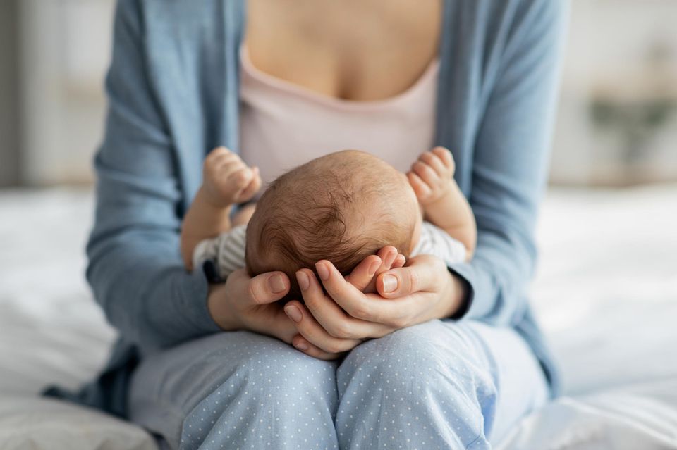 Plagiocephalus: Frau hält den Kopf eines Babys das auf ihren Knien liegt in ihren Händen