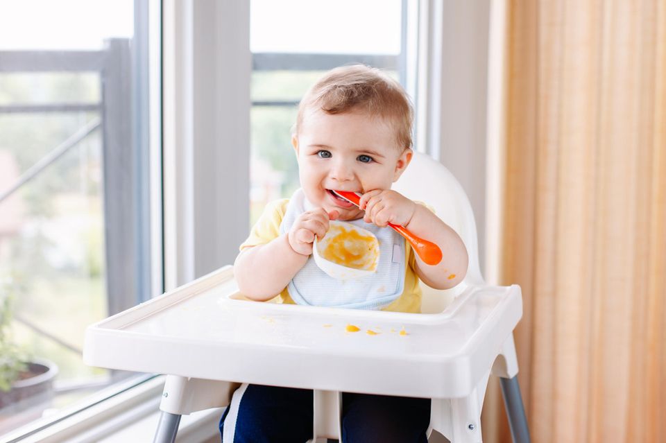 Ab wann sitzen Babys im Hochstuhl? Baby sitzt im Hochstuhl und hat einen Löffelstil im Mund