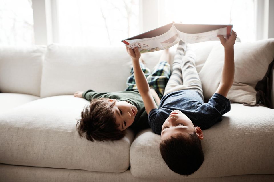 Langeweile: Zwei Kinder liegen mit dem Kopf über die Sofakante und schauen ein Buch an